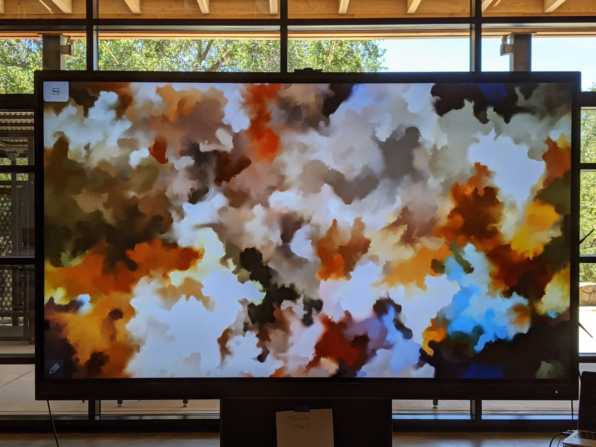 A large touchscreen on a stand in a daylit space with timber beams and a glass wall onto trees. The screen fills with soft cloud-shaped fields of orange, white, blue and brown. A small handwritten card is taped to the frame below it.