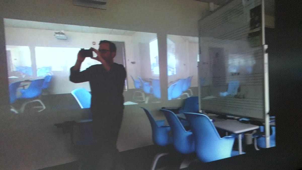 A dim classroom with a person standing in the middle holding a camera up. A projection of the same classroom falls across them and the walls, so projected chairs and windows sit behind the real blue chairs in the foreground.