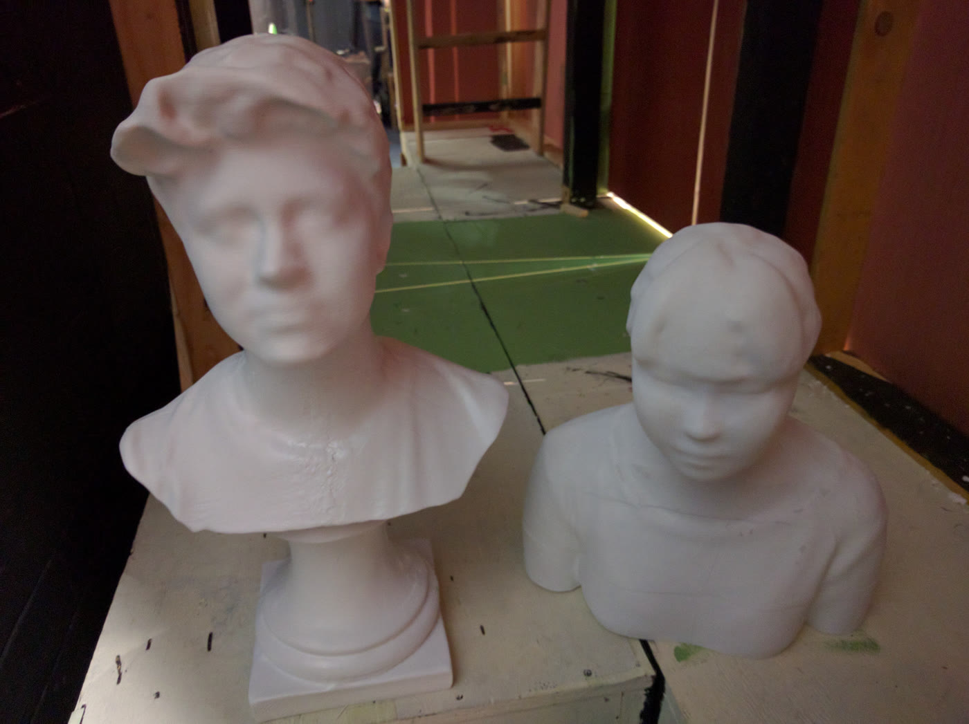 Two white 3D-printed classical busts on a workbench backstage, one on a turned plinth and one cut at the shoulders, with painted flats and a green floor behind them.