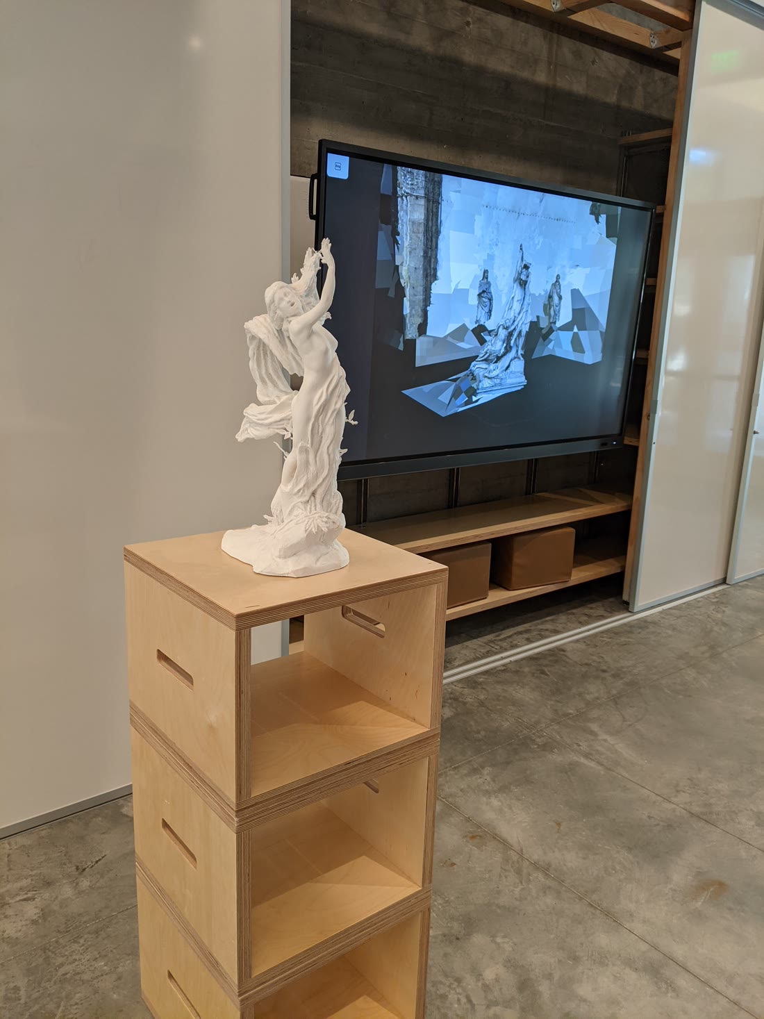 A white 3D-printed sculpture of Daphne alone, mid-transformation with drapery and laurel leaves, standing on a stacked plywood plinth in a concrete lobby. On the wall behind it a large screen displays a photogrammetric reconstruction of a Baroque sculpture group broken into pale floating plates.