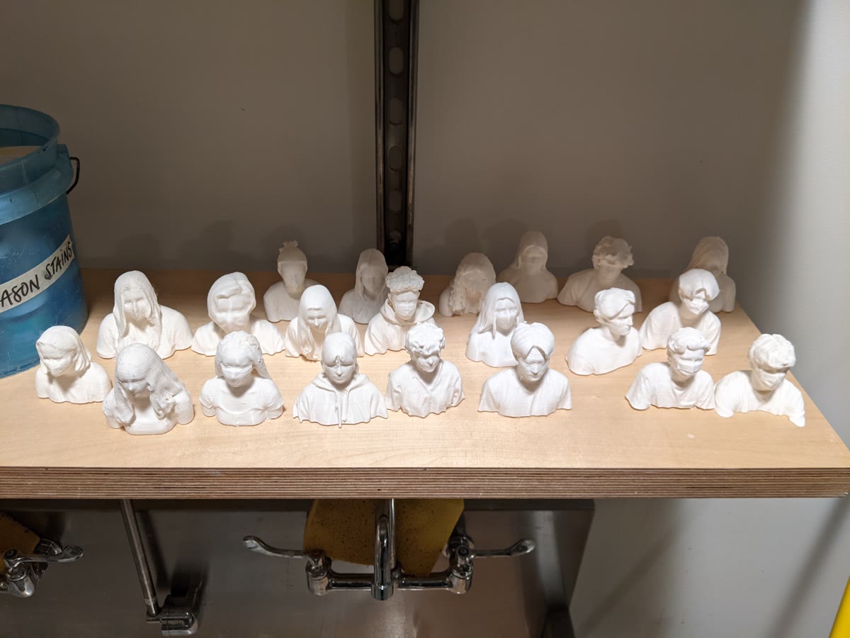 Roughly two dozen white 3D-printed portrait busts arranged in rows on a plywood shelf above a sink in a makerspace. Each one is a different student, all cleaned and finished.
